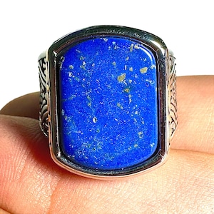 May include: A silver ring featuring a large, rectangular, deep blue lapis lazuli stone with gold flecks. The ring has an ornate, engraved design on the sides and a polished silver bezel.