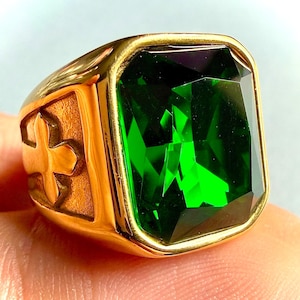 May include: A gold-toned ring featuring a large, rectangular, faceted emerald green stone set in a polished metal frame. The side of the ring band displays a raised, textured cross emblem, providing a distinct contrast to the smooth, reflective gold-colored surface of the main setting.