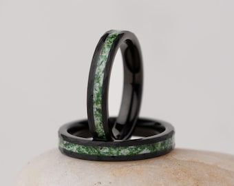 Könnte beinhalten: Zwei schwarze Ringe mit grün gesprenkelter Einlage werden auf einem glatten, hellfarbigen Stein präsentiert. Die Ringe sind gestapelt, einer aufrecht und der andere flach, um ihr Design zu zeigen. Der Hintergrund ist einfarbig neutral.