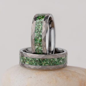 Puede incluir: Dos anillos plateados con textura martillada y una banda de piedras verdes. Un anillo está de pie, y el otro está plano sobre una pequeña piedra blanca y lisa. El fondo es de color blanco roto.