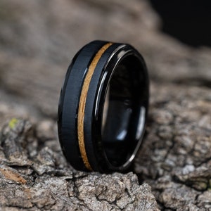 May include: A black tungsten carbide ring with a thin band of wood inlay. The ring is on a rough, textured surface.