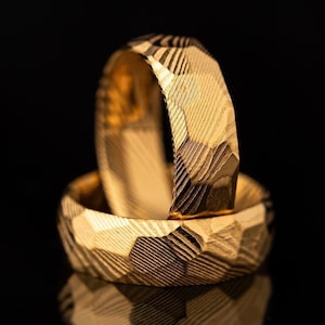 May include: Two gold wedding bands with a geometric textured pattern are stacked on a reflective black surface. The rings are in focus, showcasing the intricate design and the warm, golden hue of the metal. The background is a dark, solid colour.