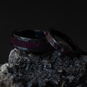 Puede incluir: Dos anillos negros con acabado texturizado e incrustaciones rojo oscuro descansan sobre una roca oscura y texturizada. Los anillos, posiblemente alianzas, se muestran sobre un fondo negro.