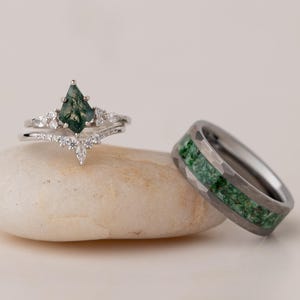 Puede incluir: Dos anillos, uno de compromiso de plata con una gema verde en forma de pera y detalles de diamantes, y otro una alianza de plata con incrustaciones verdes. Los anillos se exhiben sobre una piedra pequeña, lisa y de color claro.