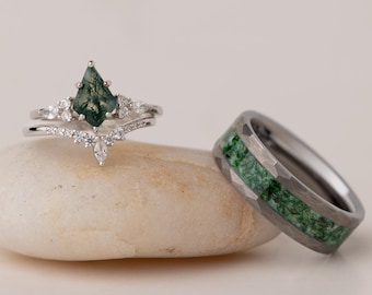 May include: Two rings, one a silver engagement ring with a pear-shaped green gemstone and diamond accents, the other a silver band with green inlay. The rings are displayed on a small, smooth, light-colored stone.