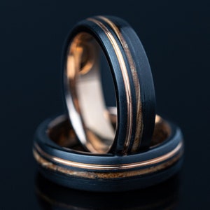May include: Two black wedding rings with a gold-colored interior and a thin gold stripe. The rings are stacked on top of each other, reflecting on a dark surface. The rings have a modern, minimalist design.