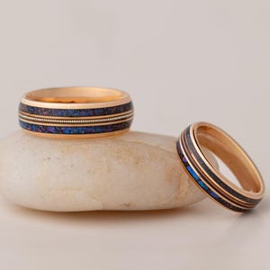 May include: Two gold-colored rings with a dark blue and gold inlay. One ring rests on a white stone, while the other is positioned to the side. The rings have a band of dark blue and gold. The rings are likely wedding bands.