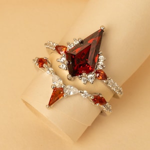 May include: Two rings with a silver band and red and white gemstones. The larger ring features a large, triangular red gemstone surrounded by smaller white gemstones. The second ring has a similar design with smaller red and white gemstones.
