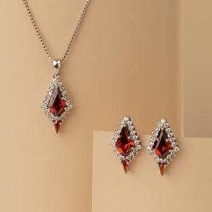 May include: A silver necklace with a pendant and matching earrings. The pendant and earrings feature a red gemstone surrounded by small white stones. The jewellery is set against a neutral background.