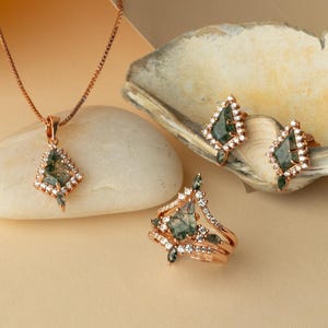 May include: A jewelry set featuring a necklace, ring, and earrings. The pieces have a rose gold finish and are adorned with green gemstones and small white accent stones. The necklace pendant and earrings are diamond-shaped, while the ring has a unique design.