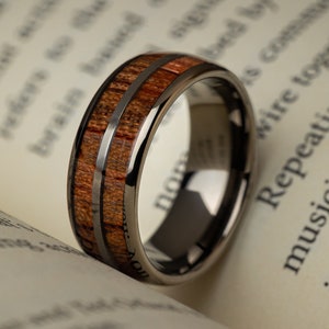 K&ouml;nnte beinhalten: Ein schwarzer Ring aus Wolframcarbid mit einer braunen Holzeinlage. Der Ring hat zwei d&uuml;nne silberne Linien, die entlang der Holzeinlage verlaufen.