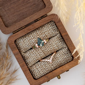 May include: Two rings in a wooden box with a burlap lining. The ring on the left has a green gemstone and small diamonds. The ring on the right is a gold band with a V-shaped row of diamonds.