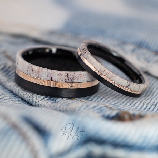 Op de afbeelding: Twee zwarte ringen van wolframcarbide met een witte antler inlay en een roségouden band. De ringen liggen op een blauwe denim achtergrond.