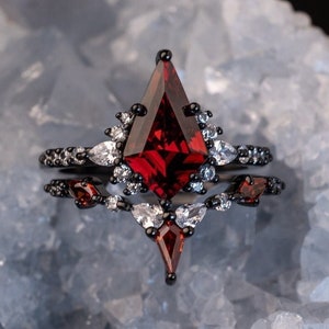 May include: A black ring with a large, red, faceted gemstone in a geometric shape. The ring is set with smaller, clear gemstones around the main stone. The ring is on a white, textured surface.