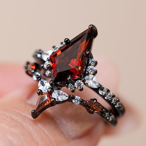 May include: A black ring set with a large, pear-shaped, red gemstone as the focal point. The ring is accented with smaller white and red gemstones. The band is black and has a delicate, ornate design. The ring is a unique piece of jewelry.