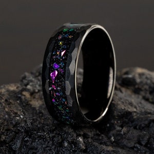 May include: A black ring with a hammered finish and a band of iridescent glitter. The ring is on a dark gray rock.