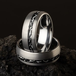 May include: Two silver wedding bands with a black inlay design. The bands are stacked on top of each other and are sitting on a dark gray rock.