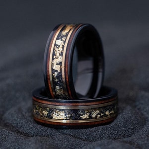 May include: Two black tungsten carbide rings with a wooden inlay and gold accents. The rings are stacked on top of each other.