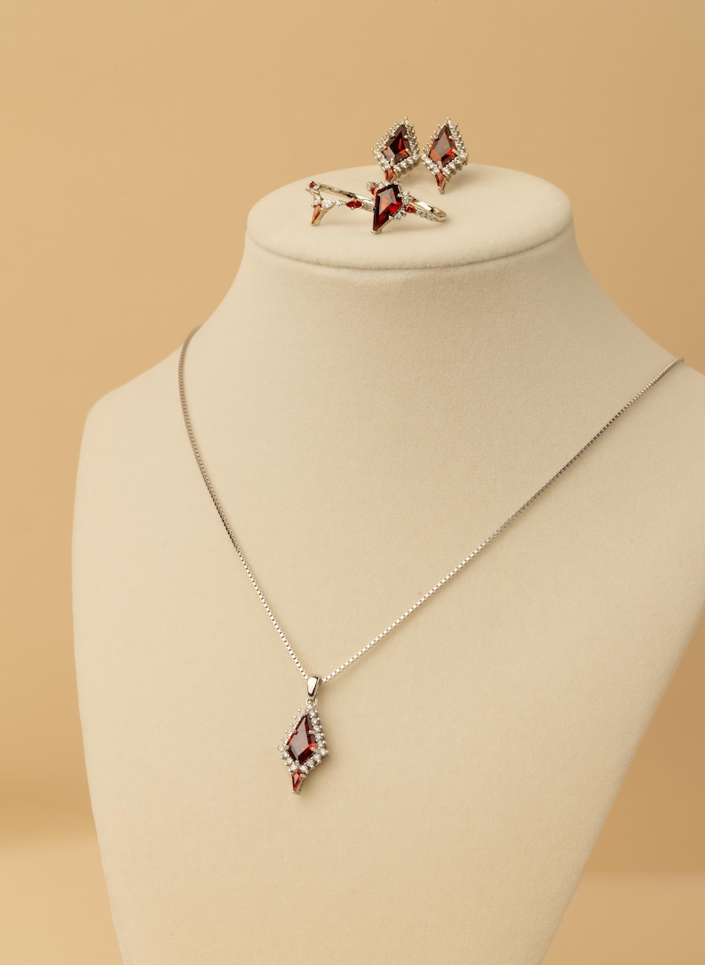 May include: A silver necklace with a red gemstone pendant, complemented by matching earrings and a ring. The jewellery features small white stones. The set is displayed on a mannequin head against a neutral backdrop. Measurements are in cm.