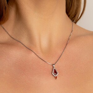 May include: A silver necklace with a diamond-shaped pendant. The pendant features a large, deep red gemstone surrounded by smaller, clear stones. The pendant hangs from a delicate chain, creating an elegant piece of jewellery.