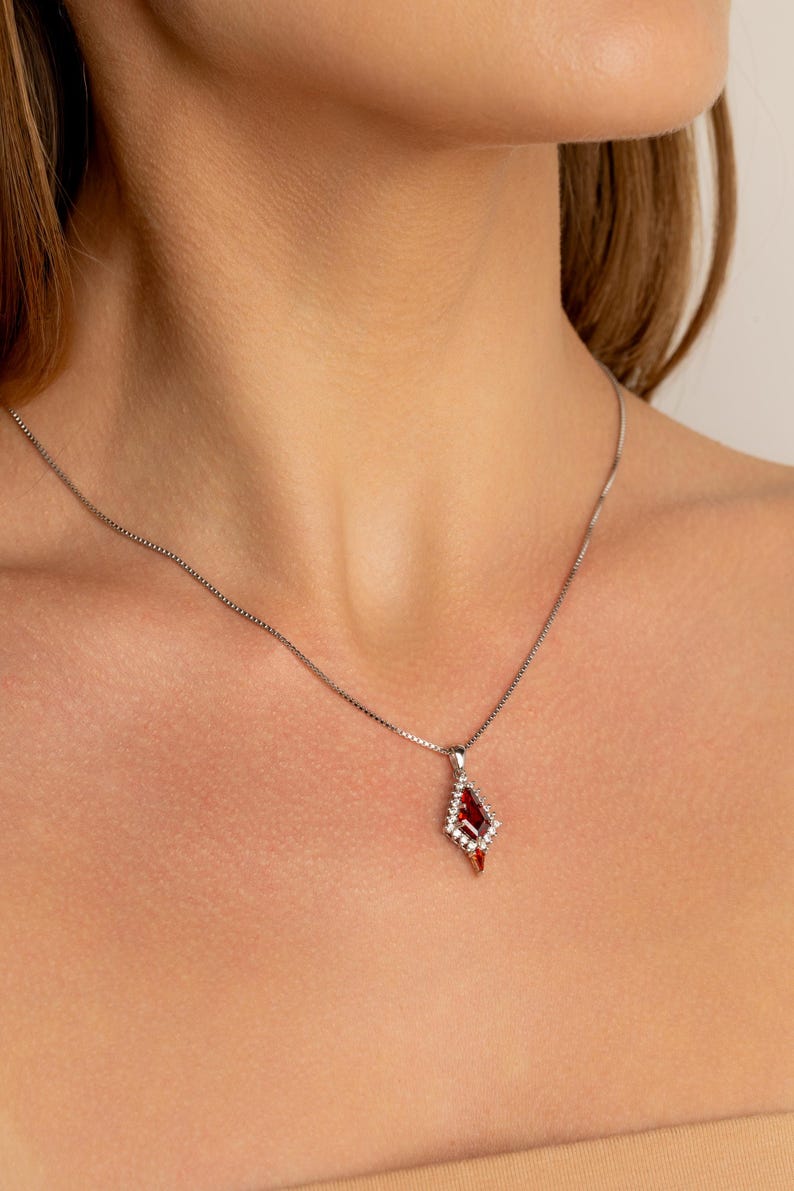 May include: A silver necklace with a diamond-shaped pendant. The pendant features a large, deep red gemstone surrounded by smaller, clear stones. The pendant hangs from a delicate chain, creating an elegant piece of jewellery.