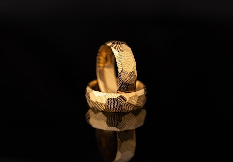 May include: Two gold wedding bands with a geometric textured pattern are stacked on a reflective black surface. The rings are in focus, showcasing the intricate design and the warm, golden hue of the metal. The background is a dark, solid colour.