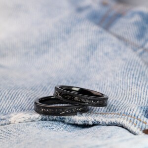 K&ouml;nnte beinhalten: Zwei schwarze Ringe mit glitzerndem Finish, &uuml;bereinander gestapelt, auf einem blauen Jeansstoff-Hintergrund.