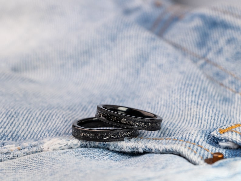 K&ouml;nnte beinhalten: Zwei schwarze Ringe mit glitzerndem Finish, &uuml;bereinander gestapelt, auf einem blauen Jeansstoff-Hintergrund.