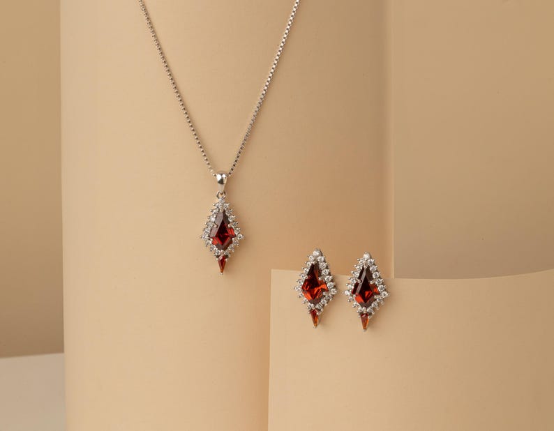 May include: A silver necklace with a pendant and matching earrings. The pendant and earrings feature a red gemstone surrounded by small white stones. The jewellery is set against a neutral background.
