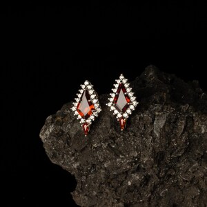 May include: Diamond-shaped earrings featuring a central red gemstone, framed by smaller white stones. The earrings are displayed on a dark, textured surface, accentuating their design and sparkle.