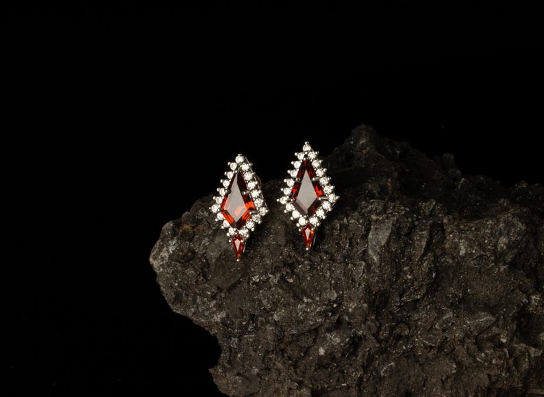 May include: Diamond-shaped earrings featuring a central red gemstone, framed by smaller white stones. The earrings are displayed on a dark, textured surface, accentuating their design and sparkle.