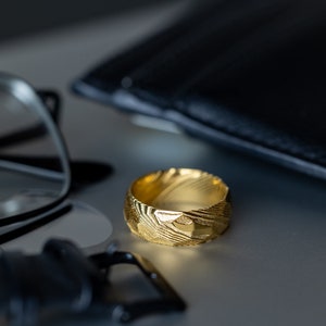 May include: A gold ring with a textured surface, set on a light surface. The ring has a pattern of lines and shapes. A black wallet and spectacles are in the background.