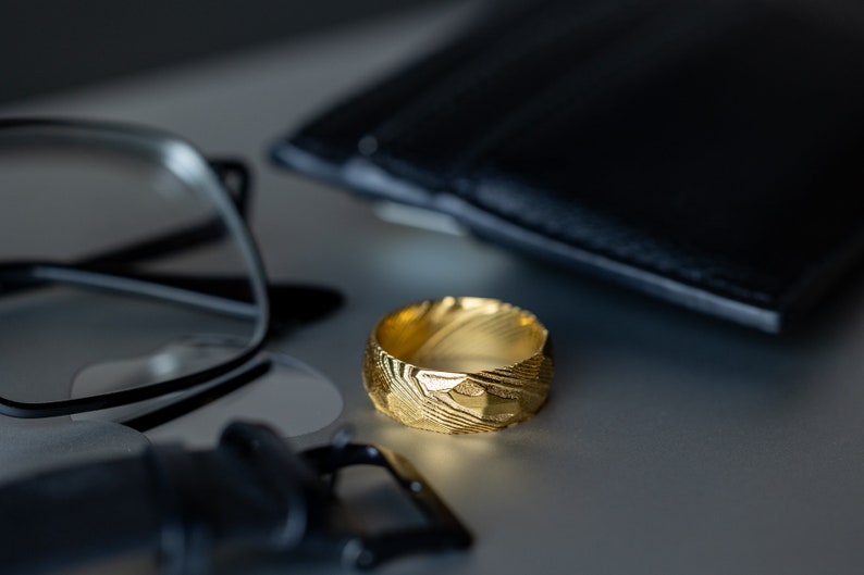 May include: A gold ring with a textured surface, set on a light surface. The ring has a pattern of lines and shapes. A black wallet and spectacles are in the background.