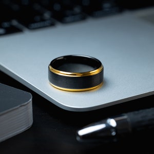 May include: A black tungsten carbide ring with a gold band. The ring is sitting on a silver laptop.