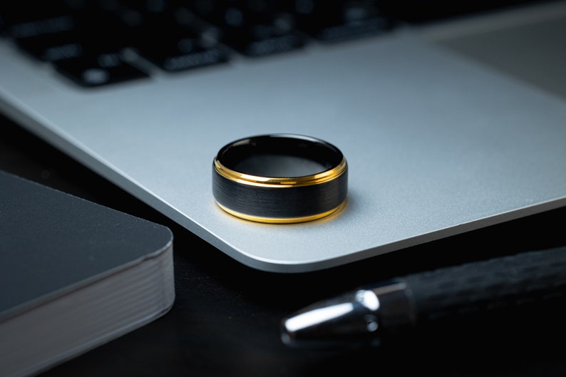 May include: A black tungsten carbide ring with a gold band. The ring is sitting on a silver laptop.