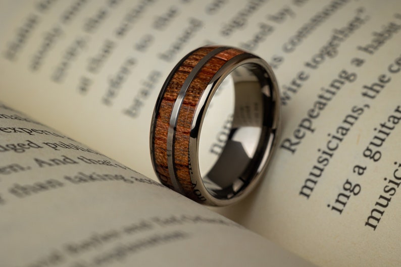 K&ouml;nnte beinhalten: Ein schwarzer Ring aus Wolframcarbid mit einer braunen Holzeinlage. Der Ring hat zwei d&uuml;nne silberne Linien, die entlang der Holzeinlage verlaufen.