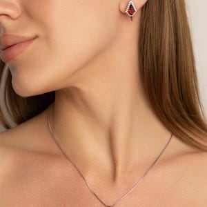 May include: A jewellery set comprising a necklace and earrings. The necklace features a silver chain with a diamond-shaped pendant with a red gemstone centre and white accent stones. The earrings mirror the pendant design. The jewellery is set against a neutral background.