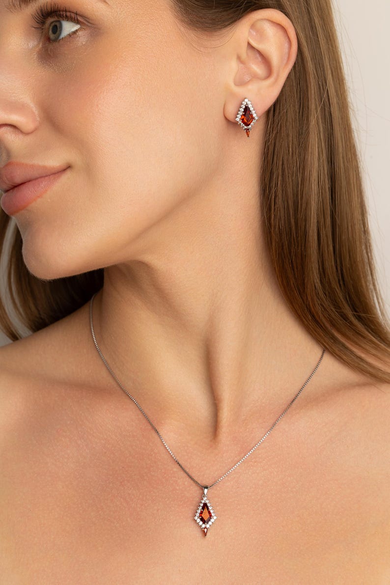 May include: A jewellery set comprising a necklace and earrings. The necklace features a silver chain with a diamond-shaped pendant with a red gemstone centre and white accent stones. The earrings mirror the pendant design. The jewellery is set against a neutral background.