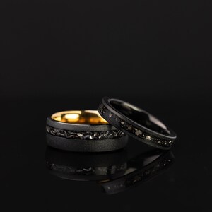 K&ouml;nnte beinhalten: Zwei schwarze Ringe mit einem d&uuml;nnen Band aus Gold und schwarzen Diamanten. Die Ringe liegen auf einer schwarzen Oberfl&auml;che.