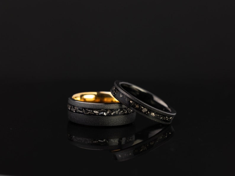 K&ouml;nnte beinhalten: Zwei schwarze Ringe mit einem d&uuml;nnen Band aus Gold und schwarzen Diamanten. Die Ringe liegen auf einer schwarzen Oberfl&auml;che.