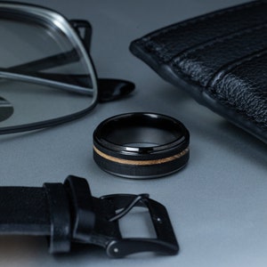May include: A black tungsten ring with a wood inlay. The ring is on a gray surface with a black leather wallet and glasses.