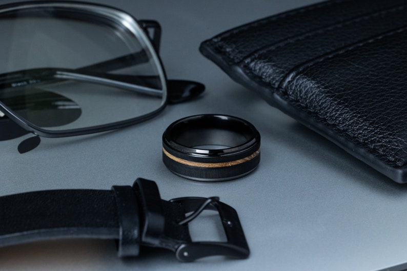 May include: A black tungsten ring with a wood inlay. The ring is on a gray surface with a black leather wallet and glasses.