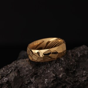 May include: A gold-coloured ring with a textured, faceted surface. The ring has a unique pattern of lines and angles, and is set against a dark background. The ring is a piece of jewellery.