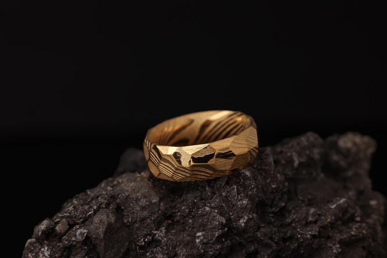 May include: A gold-coloured ring with a textured, faceted surface. The ring has a unique pattern of lines and angles, and is set against a dark background. The ring is a piece of jewellery.