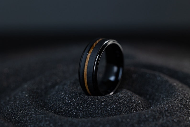 May include: A black tungsten carbide ring with a brown wood inlay. The ring is sitting on a black surface.