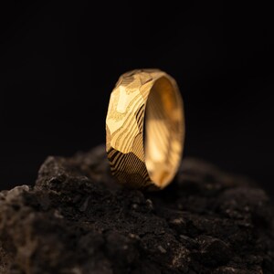 May include: A gold ring with a distinctive, textured surface. The ring is set against a dark background, emphasising its intricate design. The ring's surface has a pattern of lines and facets.