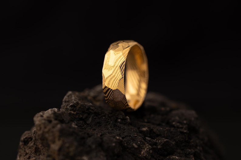 May include: A gold ring with a distinctive, textured surface. The ring is set against a dark background, emphasising its intricate design. The ring's surface has a pattern of lines and facets.