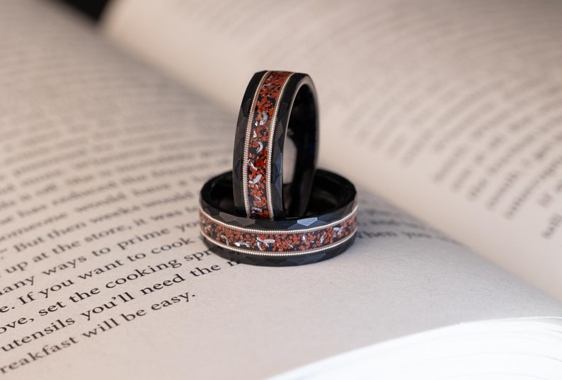 Puede incluir: Dos alianzas de boda negras y plateadas con una incrustaci&oacute;n roja y plateada. Las alianzas est&aacute;n apiladas una encima de la otra y est&aacute;n sobre un libro.