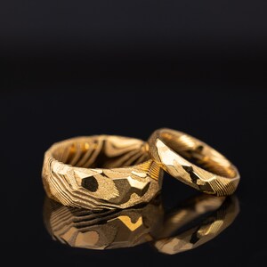 May include: Two gold wedding rings with a textured, geometric design. The larger ring has a wider band with a more pronounced pattern, while the smaller ring has a thinner band. Both rings reflect light.