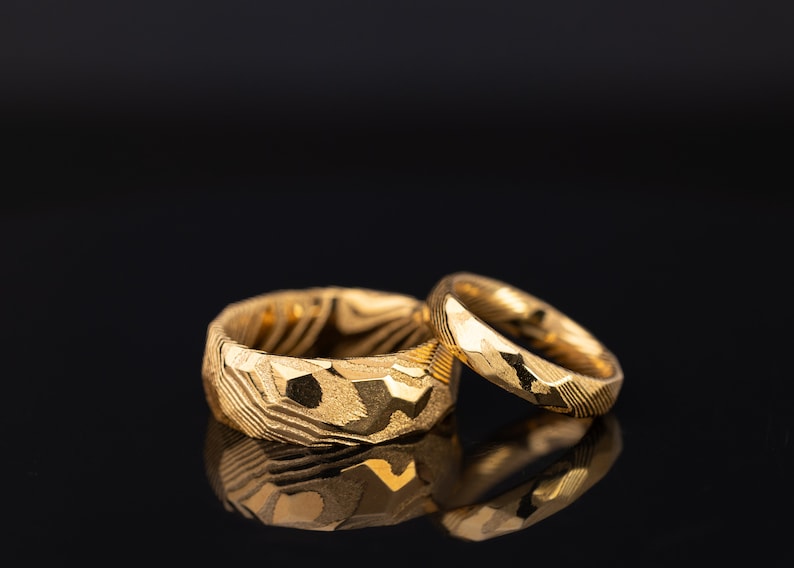 May include: Two gold wedding rings with a textured, geometric design. The larger ring has a wider band with a more pronounced pattern, while the smaller ring has a thinner band. Both rings reflect light.